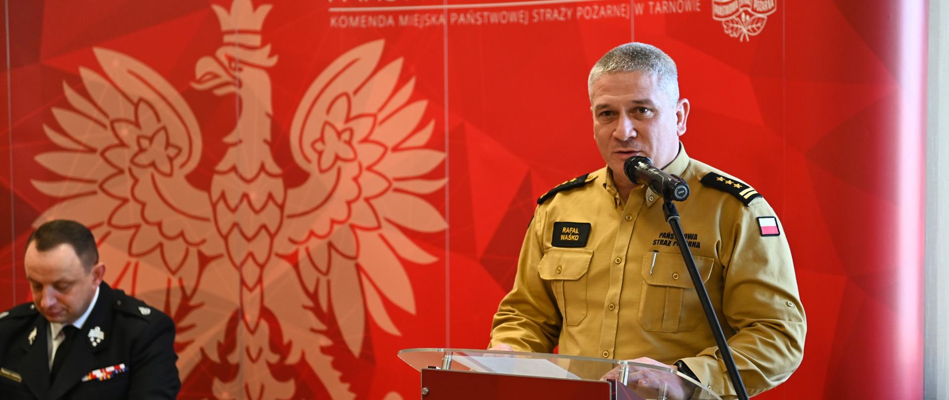 komendant ubrany w piaskowy mundur przemawia do mikrofonu z tyłu czerwony baner z logiem psp
