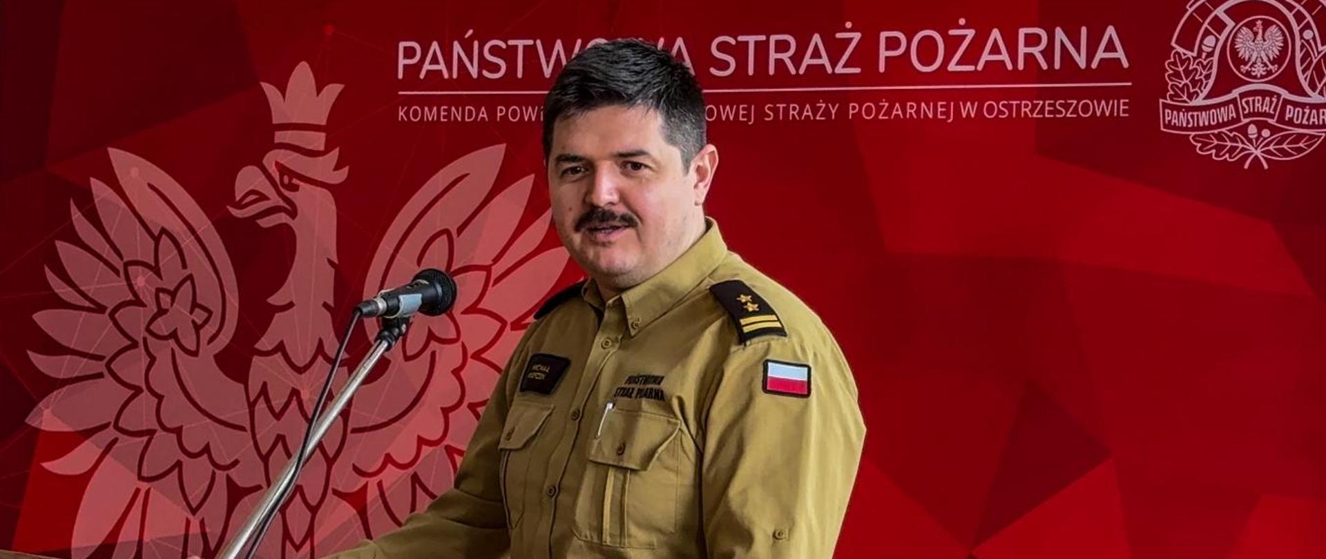Przedstawiciel Komendanta Wojewódzkiego PSP w trakcie przemówienia przed mównicą. W tle baner Państwowej Straży Pożarnej w kolorze czerwonym i z godłem Polski.