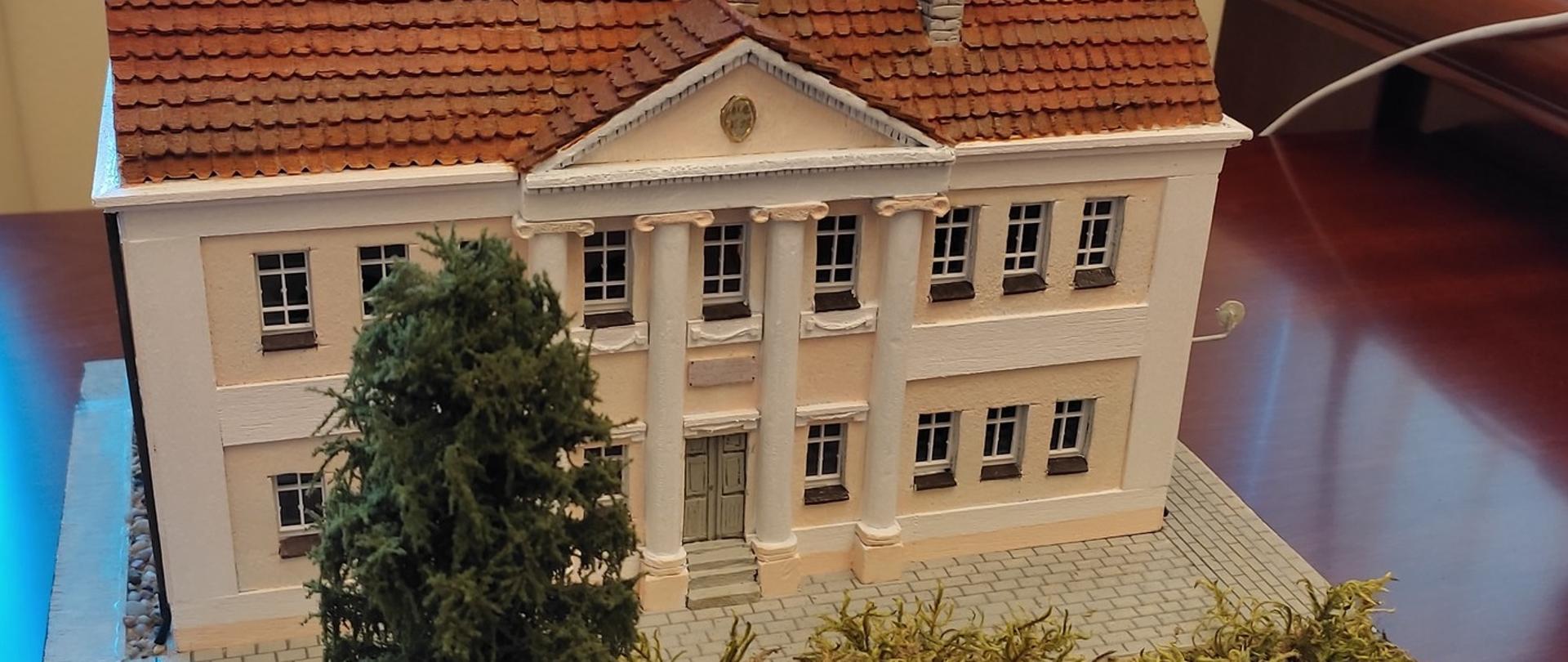 Widoczny front budynku oraz fragment placu zieleni z wysokim drzewem iglastym.