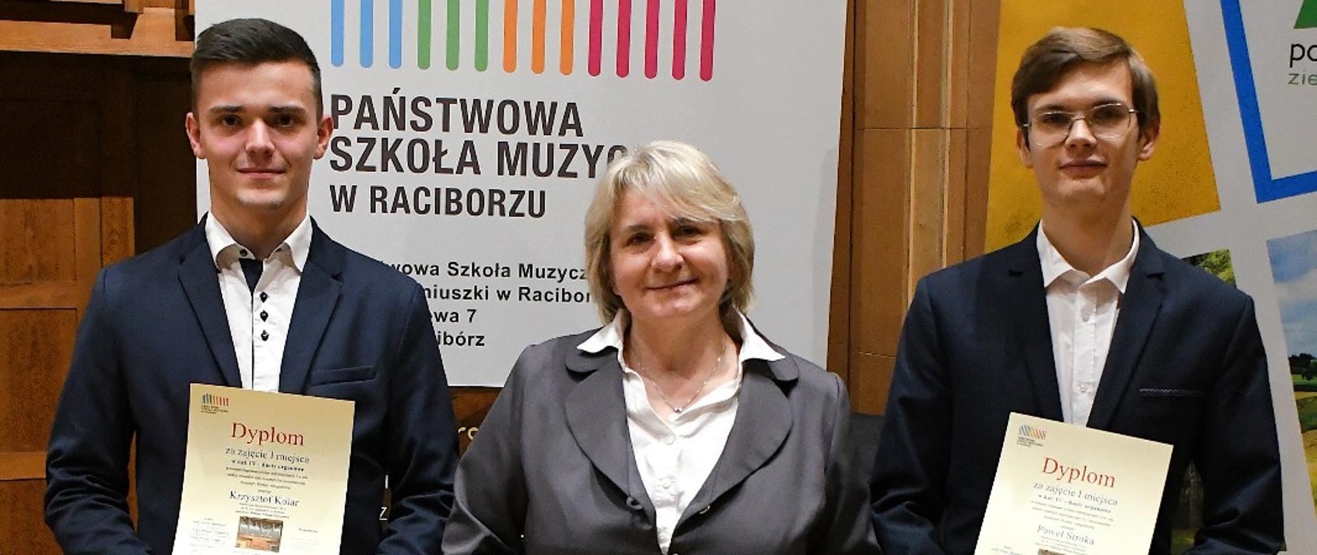 Na zdjęciu widać trzy osoby stojące w sali koncertowej w Państwowej Szkoły Muzycznej w Raciborzu. W tle znajdują się banery reklamowy tej szkoły, fragment organów i drewniane panele akustyczne.
Osoby na zdjęciu są elegancko ubrane — dwaj młodzi mężczyźni w ciemnych garniturach i w środku kobieta w szarym kostiumie. Każda z nich trzyma w rękach dyplom oraz torbę z upominkiem, co jest wynikiem zajętych miejsc podczas Transgranicznego Konkursu Organowego w Raciborzu.