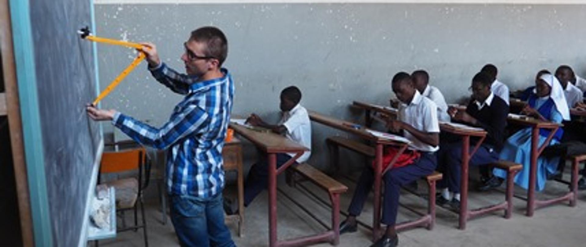 Wolontariusz rysuje cyrklem na tablicy i uczy grupę uczniów siedzących w ławkach rysunku technicznego