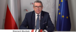 Minister Wojciech Murdzek siedzi za stołem, za nim flagi Polski i UE.