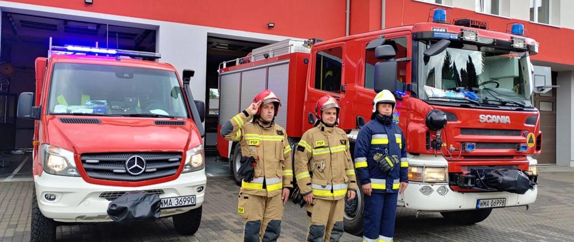 Strażacy stoją na placu przed samochodami strażackimi z włączonymi sygnałami świetnymi i dźwiękowymi w celu upamiętnienia 77. rocznicy wybuchu Powstania Warszawskiego.