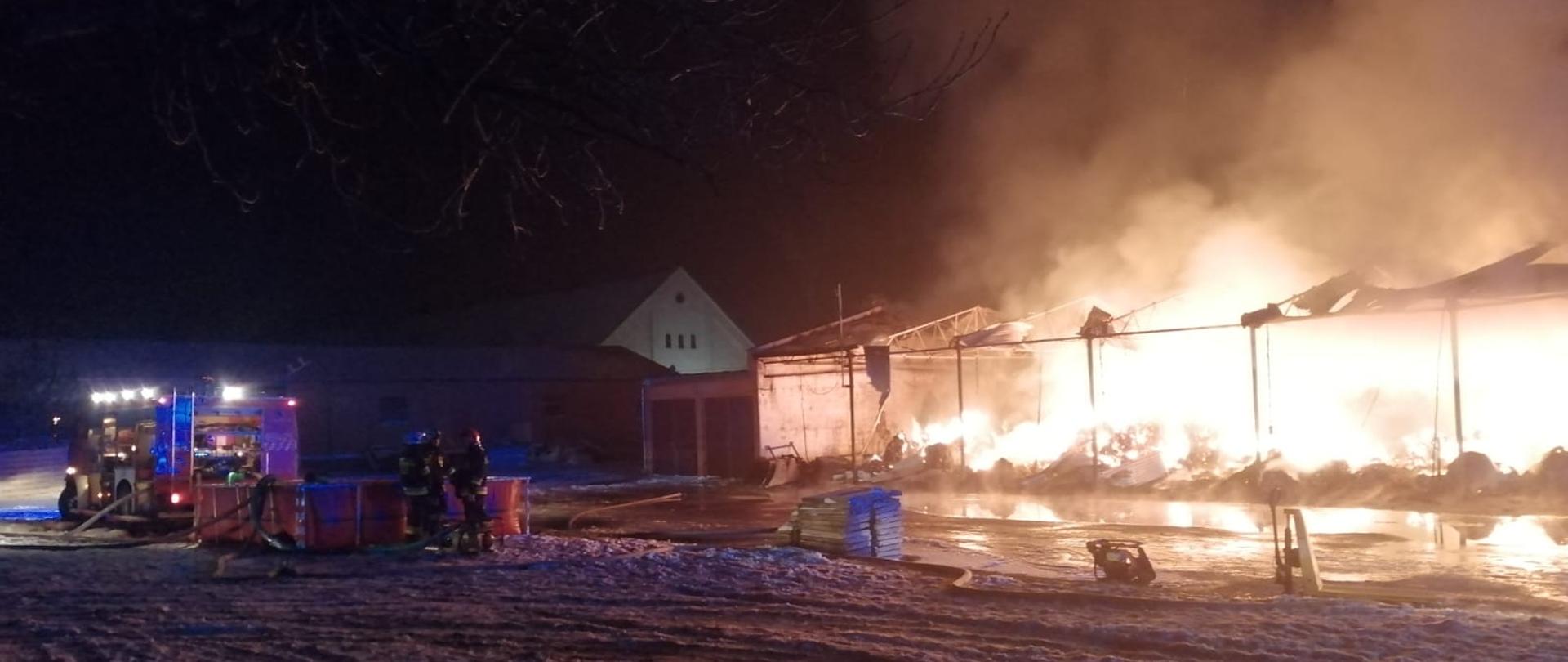 Pożar Piotrkowice. Palące się baloty siana, zawalony dach wiaty pod którą były składowane, strażacy przy samochodzie, zbiornik z zapasem wody.