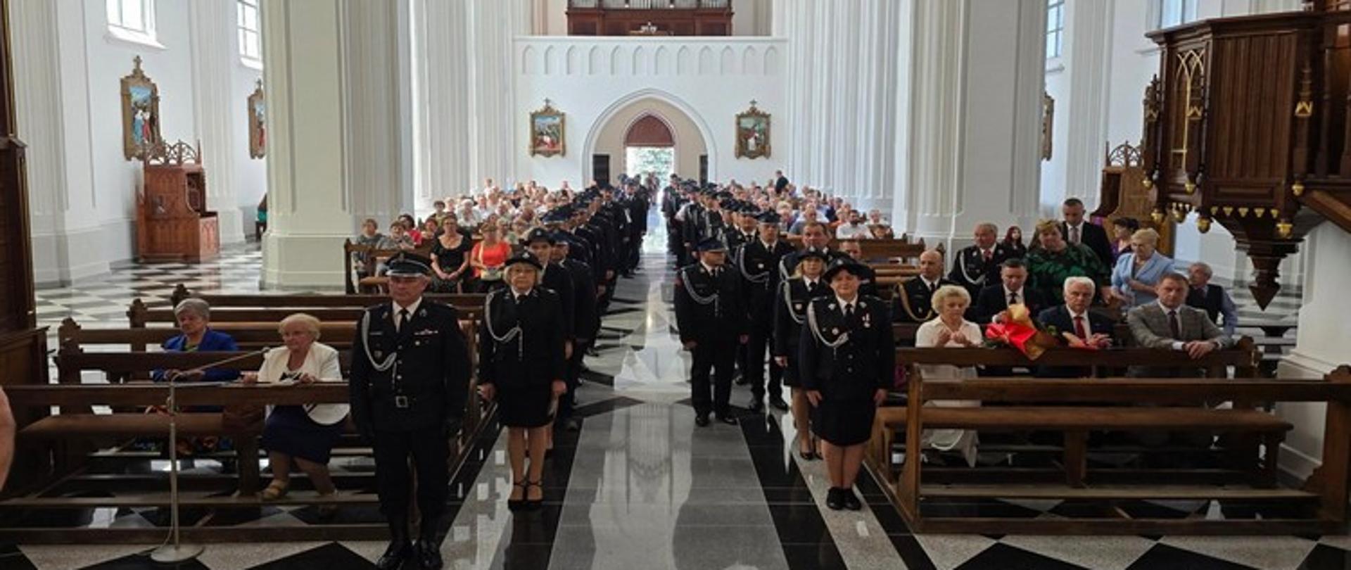 Obraz przedstawia wnętrze dużego kościoła, gdzie odbywa się uroczystość. Główną nawę wypełnia długa kolejka mężczyzn i kobiet w ciemnych, galowych mundurach, prawdopodobnie strażackich, z widocznymi odznaczeniami. Część z nich stoi, inni siedzą w ławkach. Po obu stronach nawy, w ławkach, siedzą liczni goście i wierni, obserwujący wydarzenie. Wnętrze kościoła charakteryzuje się wysokimi sklepieniami, rzędami kolumn i łukowatymi oknami. W tle widać ołtarz i inne elementy wystroju sakralnego. Podłoga jest pokryta wzorzystymi, czarno-białymi płytkami, które odbijają światło.