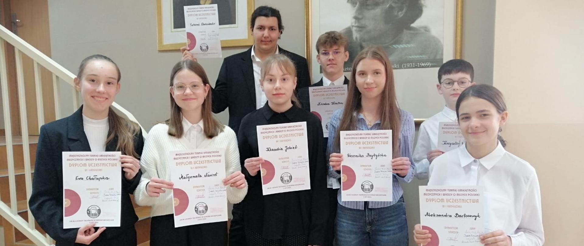 Zdjęcie przedstawia ośmioro uczniów szkoły ubranych na galowo i trzymających dyplomy na tle schodów i obrazów. 