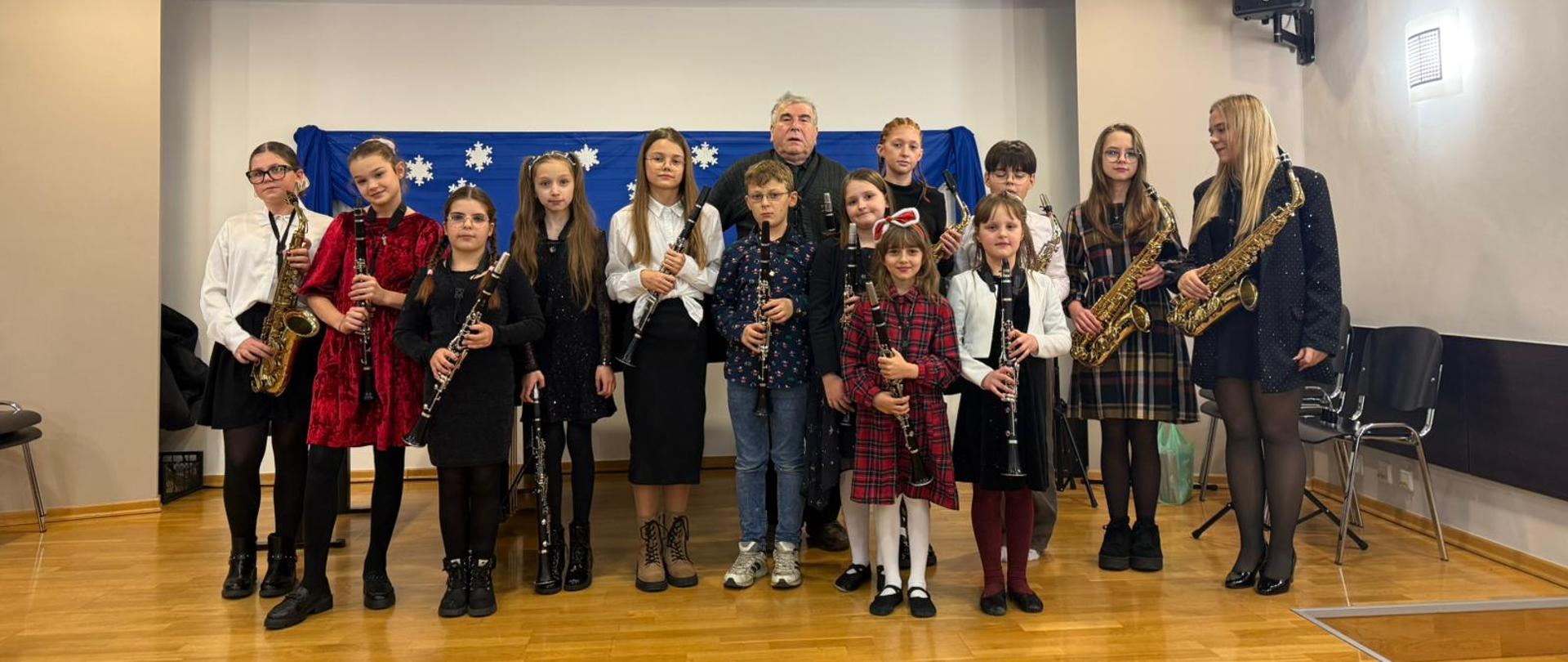 Grupa dzieci i młodzieży stoi na scenie w sali koncertowej, trzymając w rękach klarnety i saksofony. W środku grupy stoi nauczyciel. Wszyscy pozują do wspólnego zdjęcia po występie. Za nimi znajduje się niebieskie tło ozdobione białymi papierowymi śnieżynkami. Scena jest dobrze oświetlona, a uczestnicy są odświętnie ubrani.