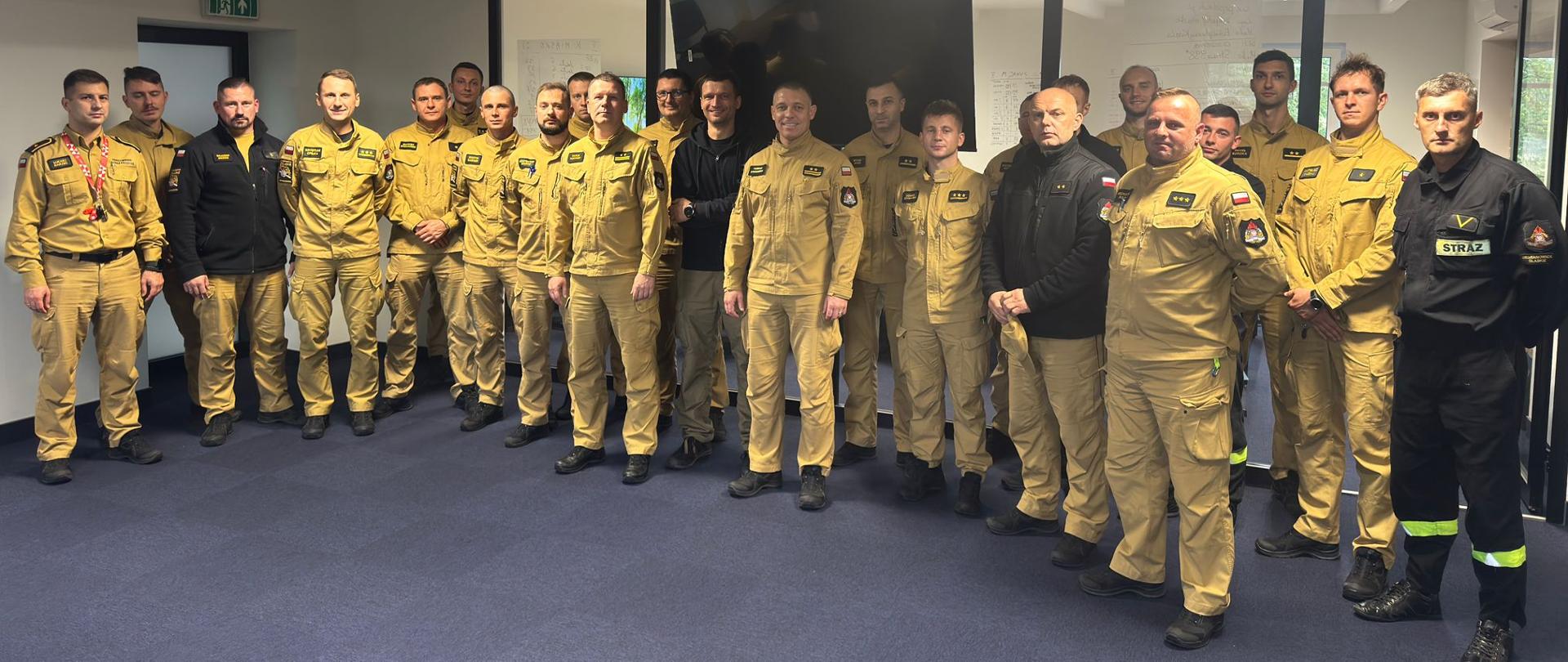Grupa około 25 funkcjonariusz państwowej straży pożarnej w mundurach służbowych koloru piaskowego i czarnego pozuje do zdjęcia grupowego w pomieszczeniu z niebieską wykładziną i białym sufitem. W tle widać szklane ściany i drzwi prowadzące do innego pomieszczenia.