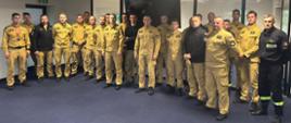 Grupa około 25 funkcjonariusz państwowej straży pożarnej w mundurach służbowych koloru piaskowego i czarnego pozuje do zdjęcia grupowego w pomieszczeniu z niebieską wykładziną i białym sufitem. W tle widać szklane ściany i drzwi prowadzące do innego pomieszczenia.