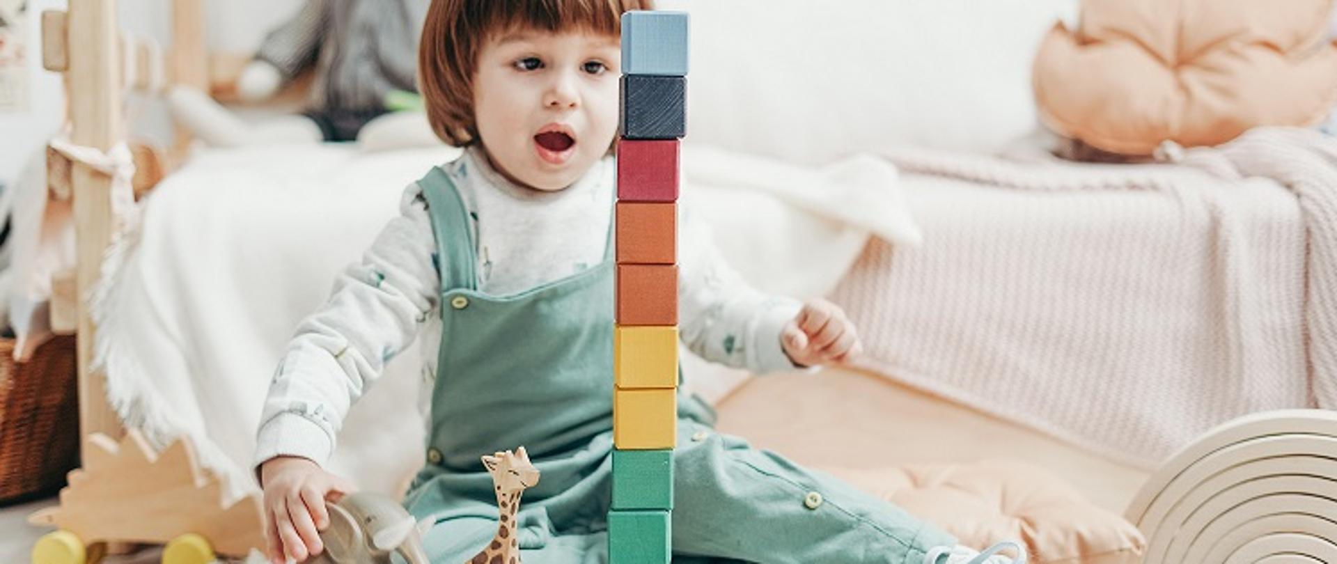 Dziecko bawiące się na podłodze drewnianymi klockami i figurkami zwierząt. 