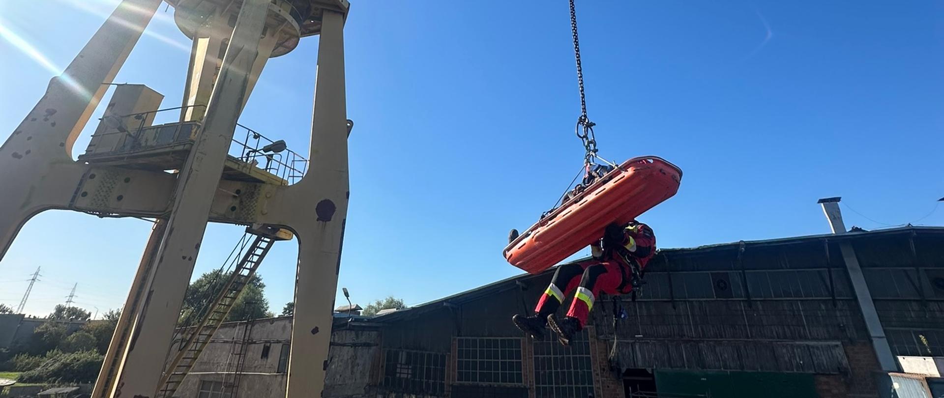 Zdjęcie przedstawia dużego, żółtego żurawia portowego, który podnosi na linie pomarańczową kapsułę ratunkową z dwoma osobami w jaskrawych kombinezonach ratowniczych. W tle widać zabudowania portowe i kilka osób stojących na ziemi, obserwujących akcję.