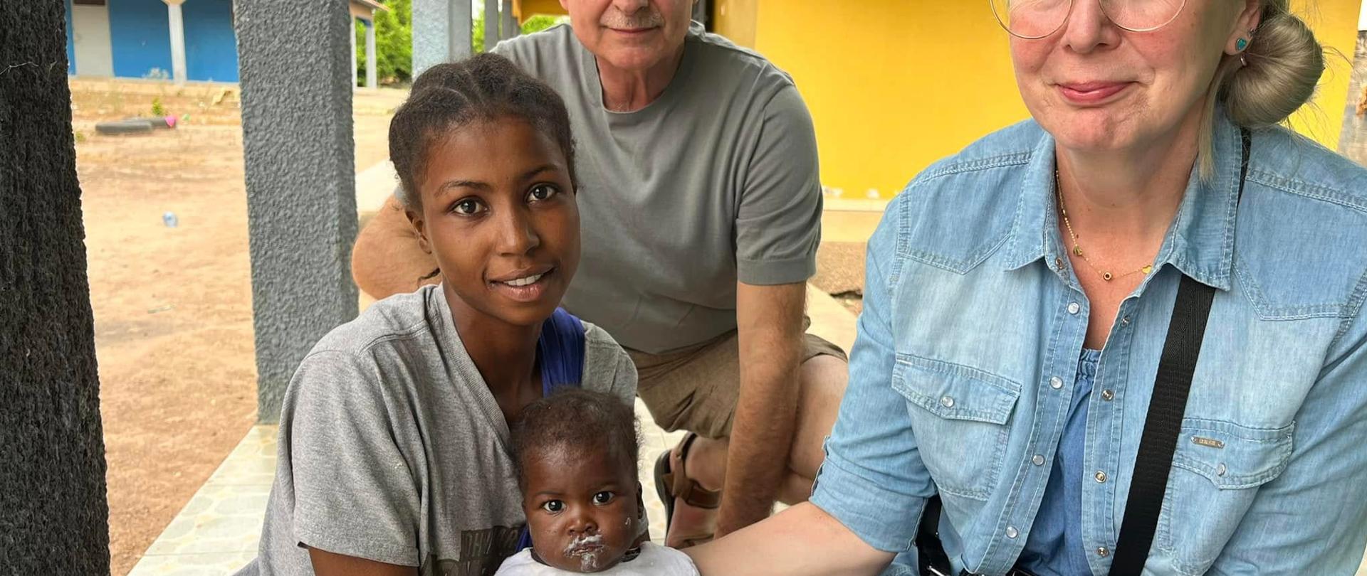 Dwie kobiety trzymają umorusane białą papką niemowlę, za nimi siedzi mężczyzna, z tyłu widać podwórko i kolorowe budynki