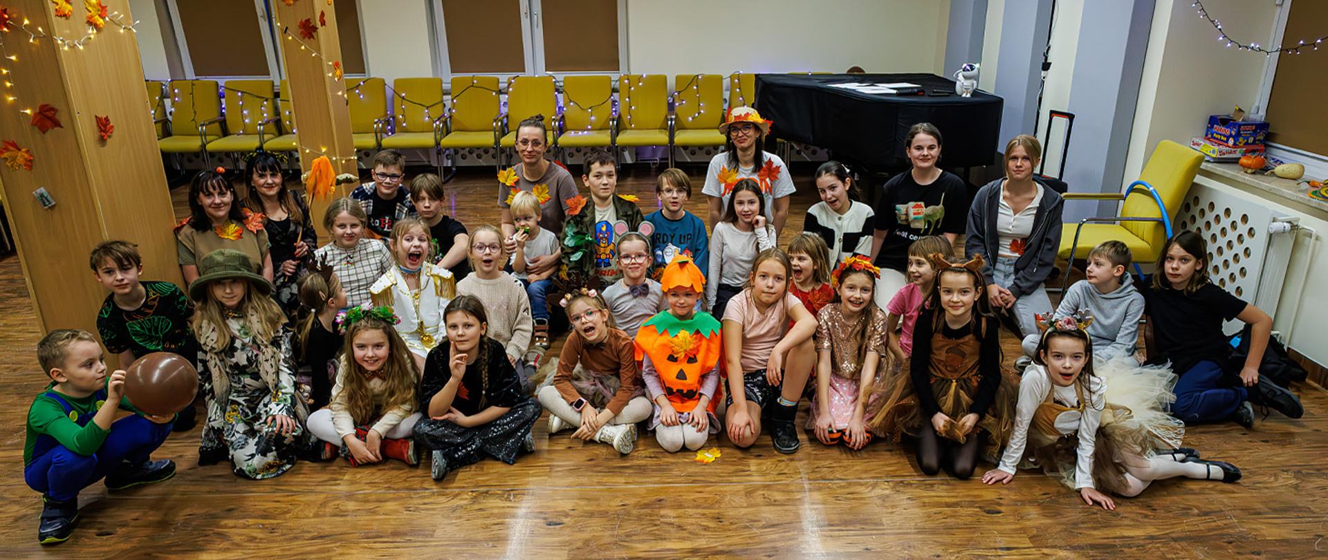 Duża grupa uczniów i nauczycieli siedzi na podłodze w auli szkolnej. Mają na sobie przebrania o tematyce jesiennej.