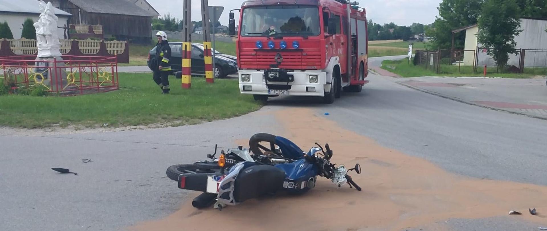 Zdjęcie przedstawia na pierwszym planie przewrócony motocykl znajdujący się na środku jezdni. Na drugim planie znajduje się samochód ratowniczo-gaśniczy oraz strażak w umundurowaniu specjalnym z białym hełmie na głowie. Wokół motocykle rozsypany sorbent neutralizujący wyciek płynów eksploatacyjnych. 