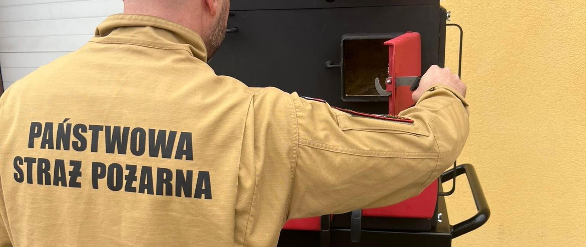 Domek i sześcian ogniowy do obserwacji zjawisk fizykochemicznych przy pożarach wewnętrznych w skali mikro - strażak obsługuje nowy sprzęt stojąc plecami do obiektywu, w tle biała brama garażowa