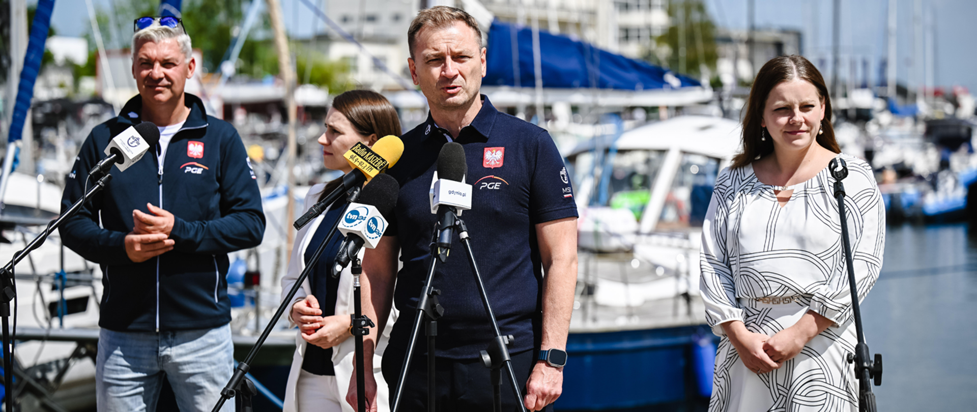 Konferencja Ministra Sportu i Turystyki Sławomira Nitrasa na świeżym powietrzu w Gdyni, 5 lipca 2024. Minister Nitras stoi za mikrofonami. Nieco z tyłu za nim od lewej strony stoją: Prezes Polskiego Związku Żeglarskiego Tomasz Chamera, posłanka Agnieszka Pomaska, Prezydentka Gdyni Aleksandra Kosiorek. W tle marina z łodziami.