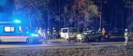 Uszkodzony samochód osobowy i bus stoją po zderzeniu na jezdni. Wokół pracujący strażacy. Obok stoi karetka pogotowia ratunkowego.