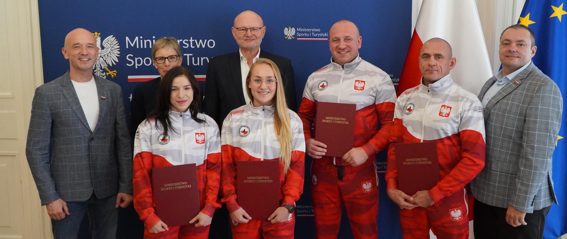 Zdjęcie zbiorowe uczestników spotkania przedstawicieli Ministerstwa Sportu i Turystyki z zawodnikami, trenerami i przedstawicielami Polskiego Związku Muaythai. Od lewej stoją: prezes Polskiego Związku Muaythai Dawid Polok, naczelnik Departamentu Sportu Wyczynowego MSiT Anna Kuder, podsekretarz stanu w MSiT Ireneusz Nalazek, trenerzy kadry narodowej Rafał Maciaszek i Maciej Domińczak oraz dyrektor ds. rozwoju i pozyskiwania funduszy Polskiego Związku Muaythai Bartłomiej Malinowski. Przed nimi zawodniczki Roksana Dargiel i Martyna Kierczyńska. Zawodniczki i trenerzy trzymają w bordowe teczki z napisem "Ministerstwo Sportu i Turystyki". Za plecami uczestników spotkania ścianka z logotypami MSiT. Z prawej strony flagi Polski i UE.