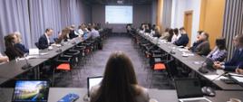 Salka konferencyjna, w której ustawione są stoły w literę U. Przy stołach siedzą uczestnicy spotkania. Na stołach stoją mikrofony oraz laptopy. W oddali wyświetlana jest prezentacja.