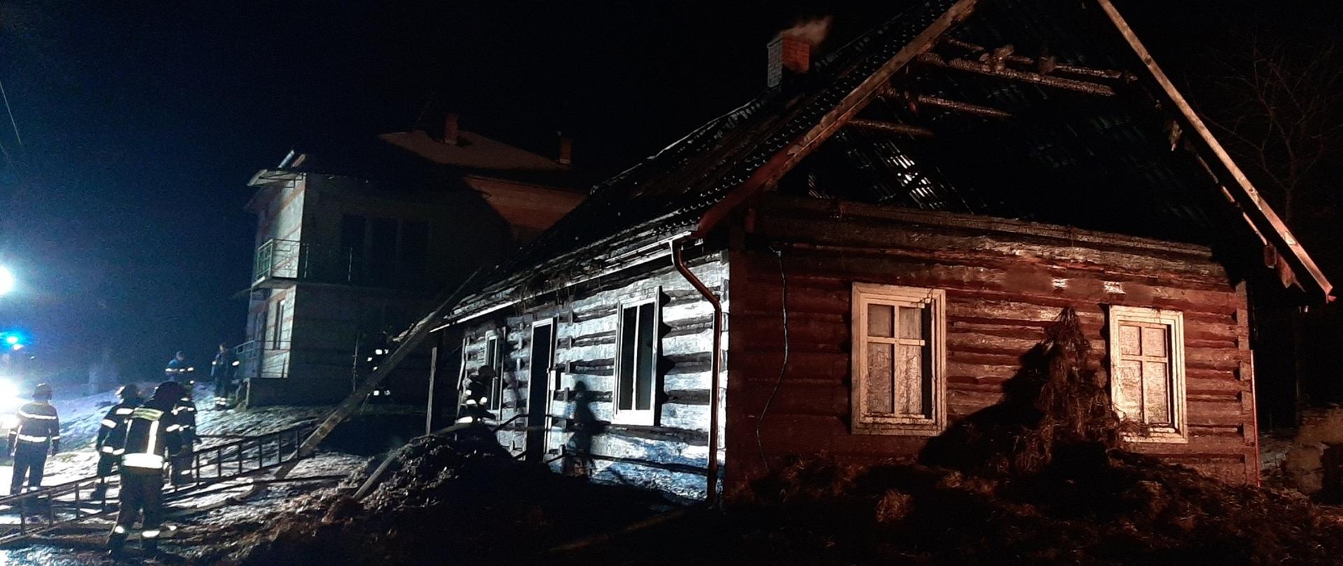 Zdjęcie zrobione na zewnątrz w nocy. Na pierwszym planie drewniany dom, o który oparta jest drabina. Po lewej stronie strażacy prowadzący działania ratownicze. W tle stoi murowany dom.