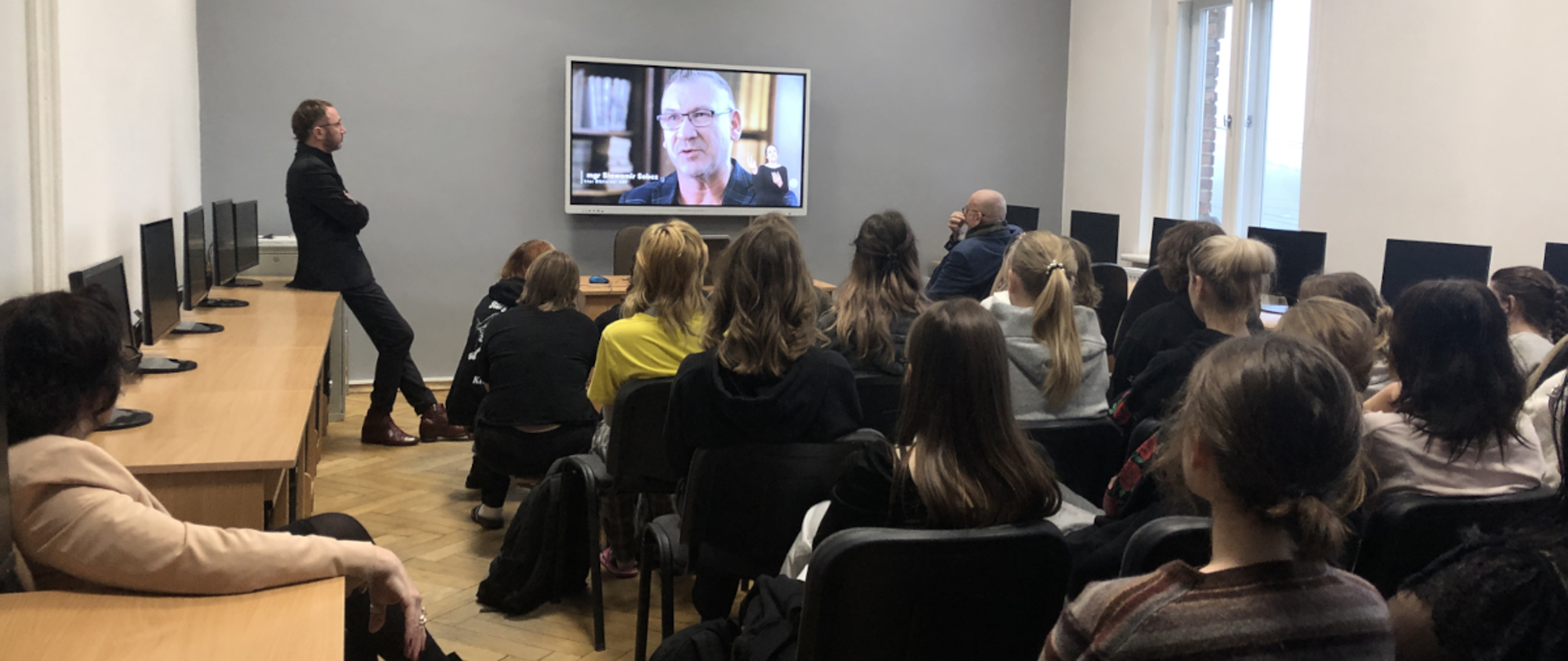 Pracownia komputerowa. Na środku siedzą tyłem słuchacze. W głębi monitor z prezentacją. Przy nim dwaj prelegenci. Jeden, z lewej przysiadł na stoliku, drugi z prawej przysłuchuje się wywiadowi prezentowanemu na monitorze.