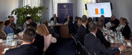 Sala konferencyjna wypełniona osobami siedzącymi przy dwóch długich stołach podczas oficjalnego spotkania. W tle widoczny ekran z prezentacja.