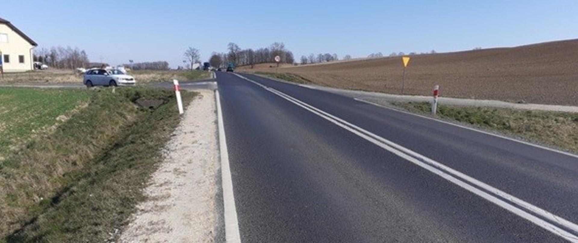 Skrzyżowanie dwóch dróg. Po lewej stronie zabudowa mieszkaniowa, po prawej pola uprawne.