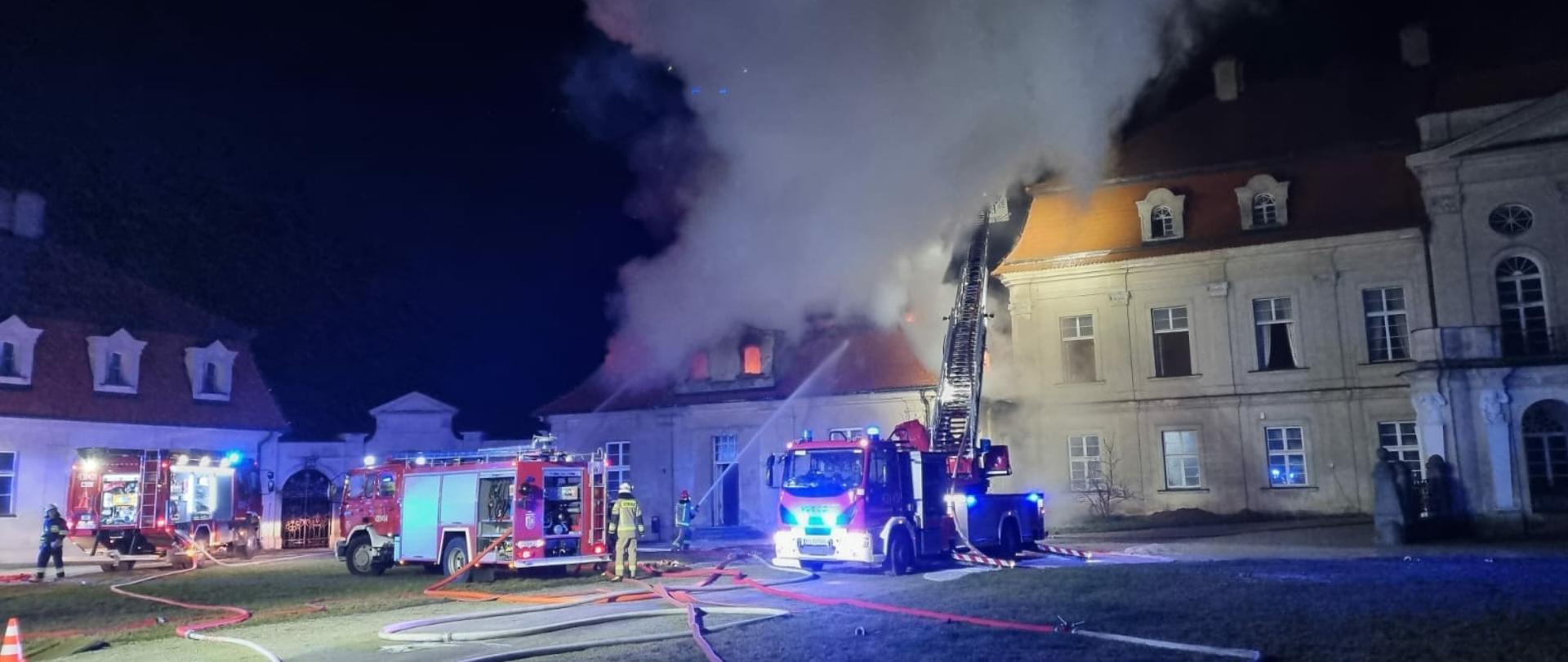 Zdjęcie przedstawia pożar alkierza przy pałacu. Pożarem objęte jest poddasze. Strażacy podają wodę na palący się dach. Na zdjęciu 3 wozy strażackie.