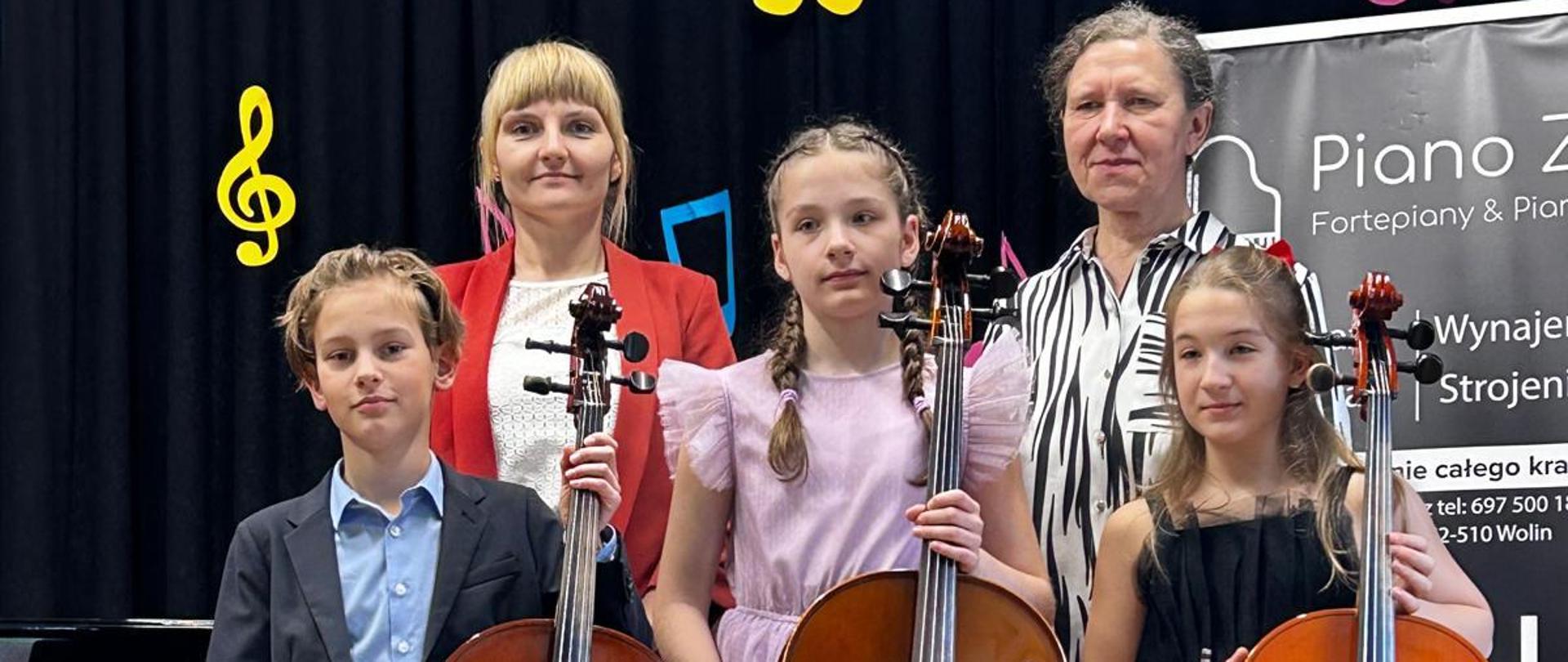 Na zdjęciu stoją 3 uczniowie z wiolonczelami oraz naucz. Dorota Zganiacz i Maria Rybak