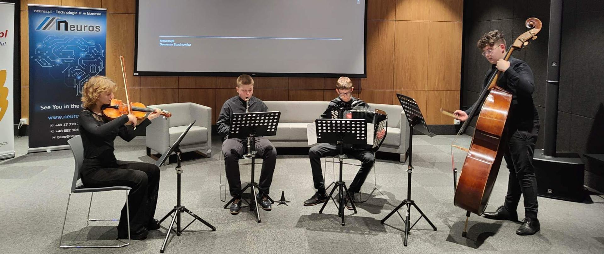 zdjęcie przedstawia członków zespołu kameralnego w składzie skrzypce, klarnet, akordeon i kontrabas podczas występu w sali kameralnej Biblioteki Miejskiej w Mielcu, zespół umieszczony jest w centralnej części zdjęcia, podłoga wyłożona jest szarą wykładziną, natomiast ściany mają czarny kolor, za muzykami widać rozsuwany ekran projekcyjny oraz obłożona boazerią ścianę