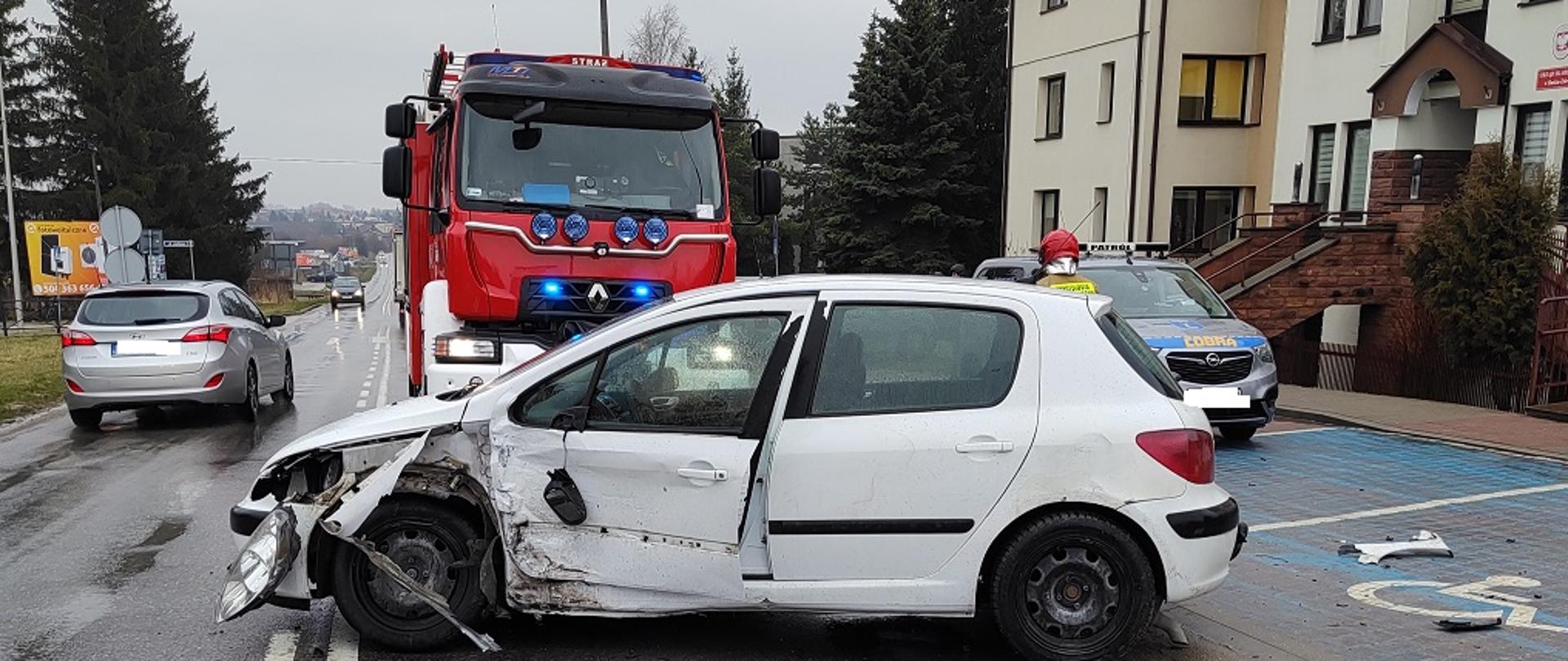Zdjęcie przedstawia uszkodzony samochód, a w tle stojący samochód pożarniczy. Po prawej stronie idzie strażak