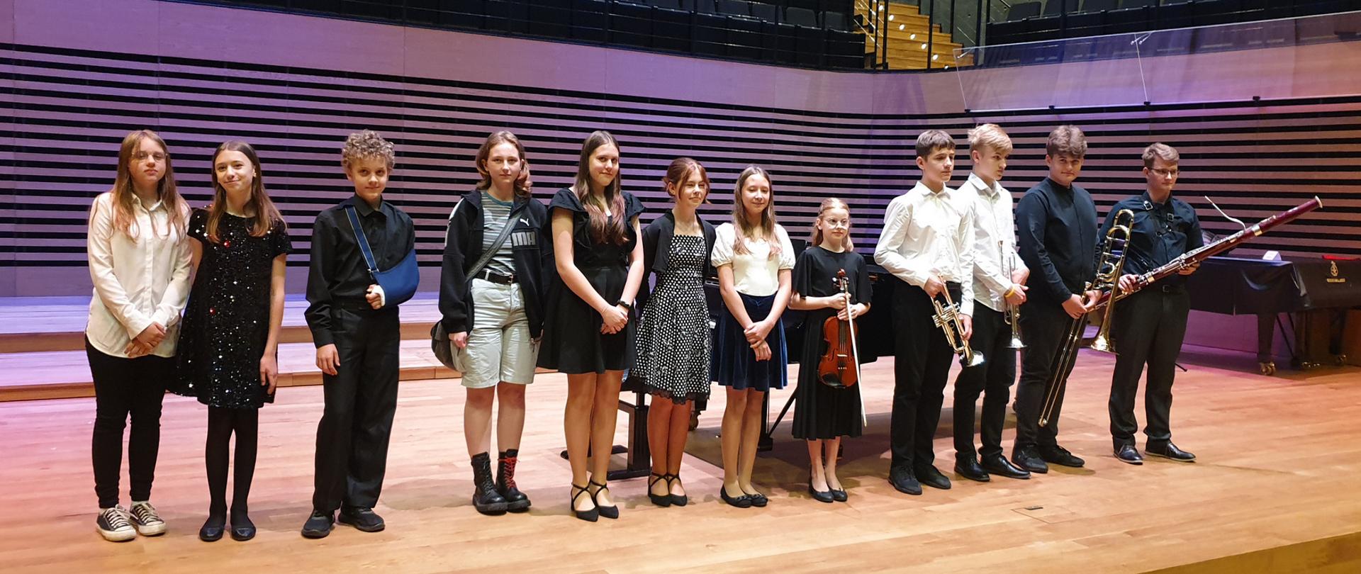 Pięciu chłopców i siedem dziewcząt stoi w rzędzie na estradzie sali koncertowej, Niektórzy trzymają przed sobą instrumenty: skrzypce, dwie trąbki, puzon i fagot.