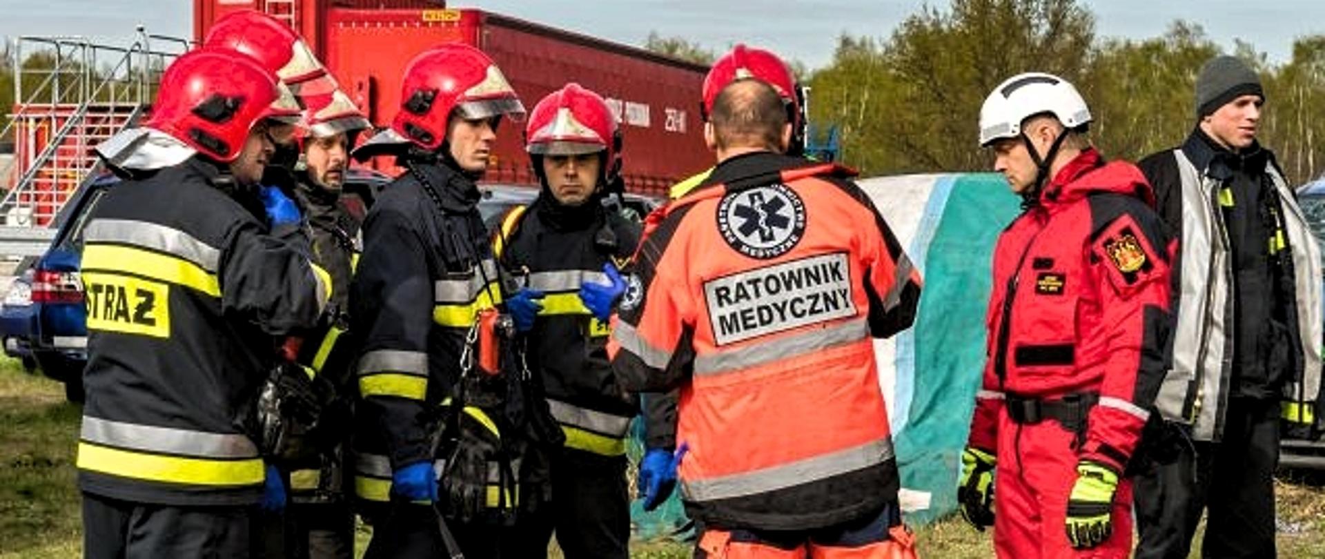 Grupa strażaków w ubraniu specjalnym i ratownik medyczny w trakcie omawiania założenia do ćwiczeń na poligonie 