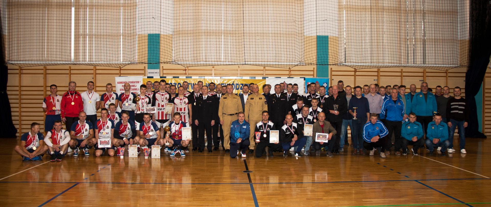 Zdjęcie przedstawia wszystkich uczestników i organizatorów finału XXIII Mistrzostw województwa podkarpackiego w piłce siatkowej ustawionych na hali sportowej w Cmolasie po zakończeniu zawodów. Zawodnicy mają przewieszone medale, w rękach trzymają dyplomy oraz puchary.