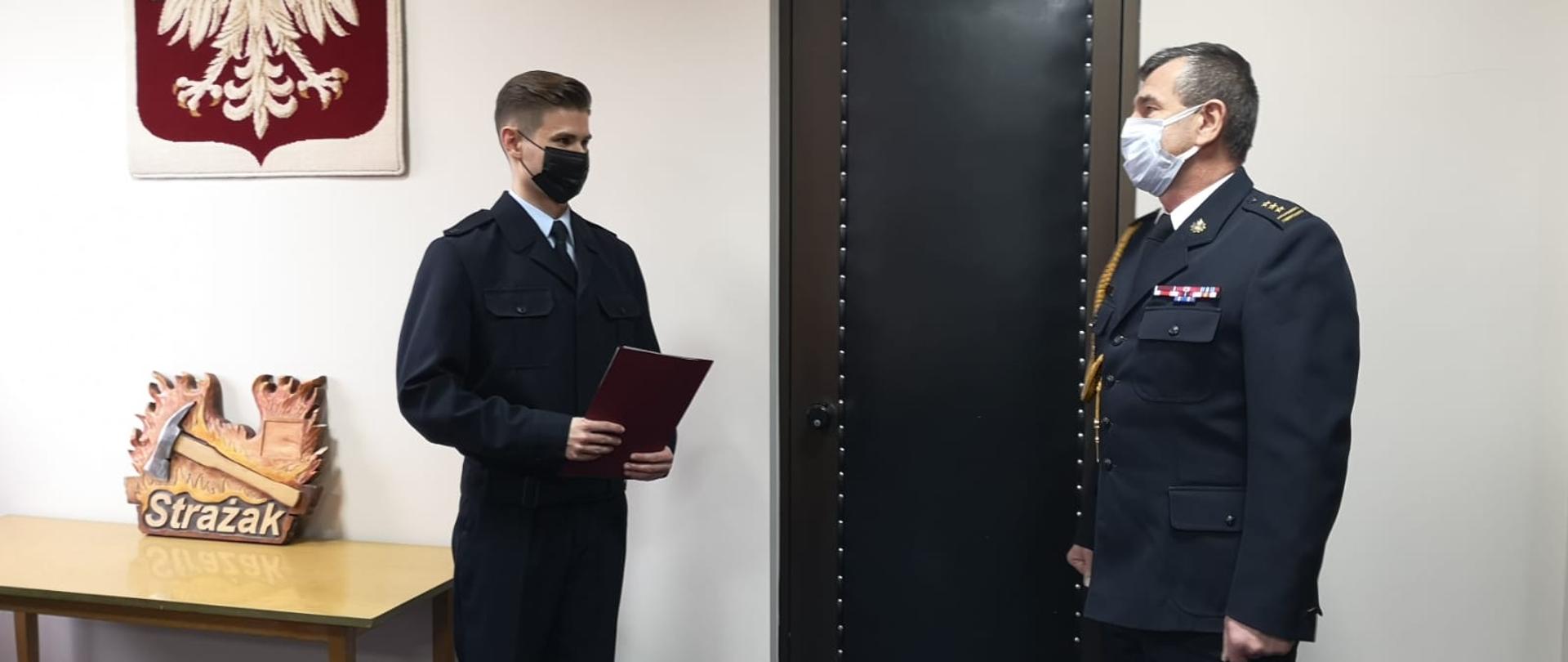 Na zdjęciu st. bryg. Janisław Buller - Komendant Miejski PSP w Bydgoszczy w mundurze wyjściowym oraz str. Filip Kukliński trzymający w dłoniach akt ślubowania. Osoby znajdują się w świetlicy budynku Komendy Miejskiej PSP w Bydgoszczy. Na tle zdjęcia kremowa ściana oraz Godło Polski.