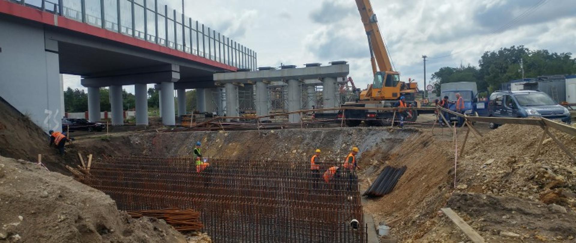 Na obrazku widoczny jest plac budowy, na którego terenie znajdują się maszyny budowlane. Widoczne są również materiały budowlane i pracownicy budowy. 
