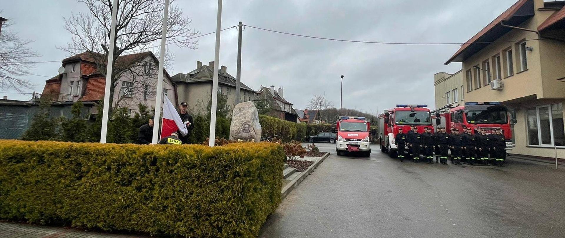 Poczet sztandarowy wciągający flagę na maszt. Z lewej strony u.stawieni w dwuszeregu strażacy przed samochodami pożarniczymi 