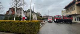 Poczet sztandarowy wciągający flagę na maszt. Z lewej strony u.stawieni w dwuszeregu strażacy przed samochodami pożarniczymi 