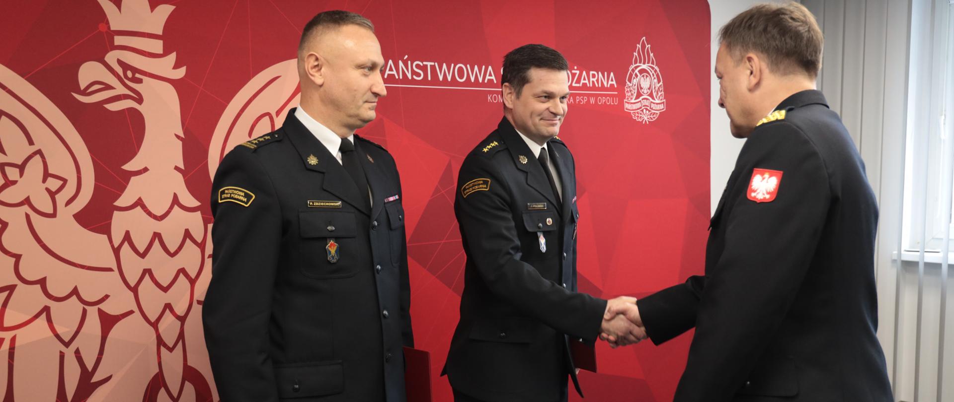 Na zdjęciu widać funkcjonariuszy w mundurach wyjściowych w pomieszczeniu podczas oficjalnej uroczystości wręczenia pochwał za za dokonanie czynu świadczącego o wyjątkowej odwadze. Dwóch strazaków stoi na tle czerwonej ścianki z wizerunkiem orła i napisem „Państwowa Straż Pożarna”, odbierając gratulacje od przełożonego w mundurze służbowym.