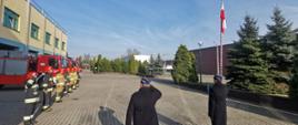 Strażacy stoją na palcu przed budynkiem strażnicy. Stoją na baczność i salutują. W tle widać budynek strażnicy przed którym ustawione są samochody strażackie. 