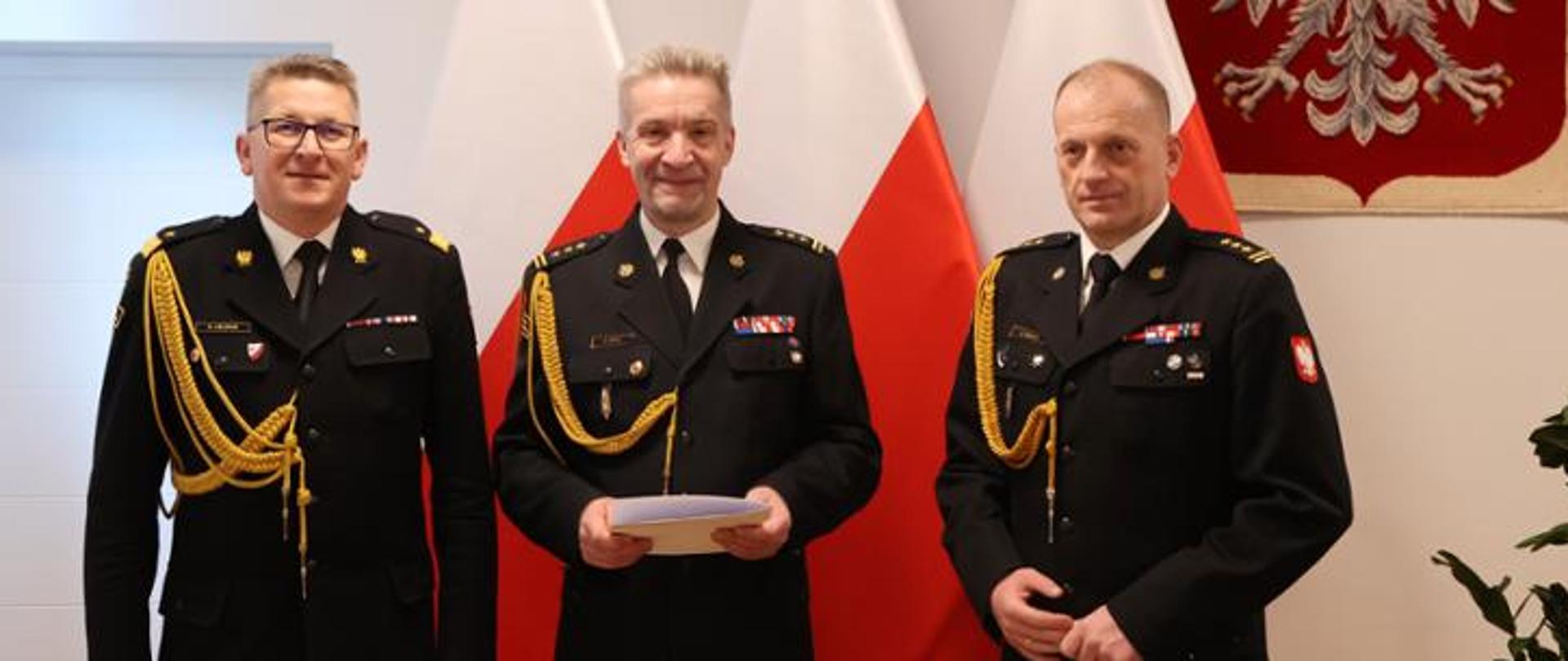 Na zdjęciu trzech funkcjonariuszy w umundurowaniu galowym ze sznurami pozuje do zdjęcia na tle biało czerwonych flag oraz godła Polski.
