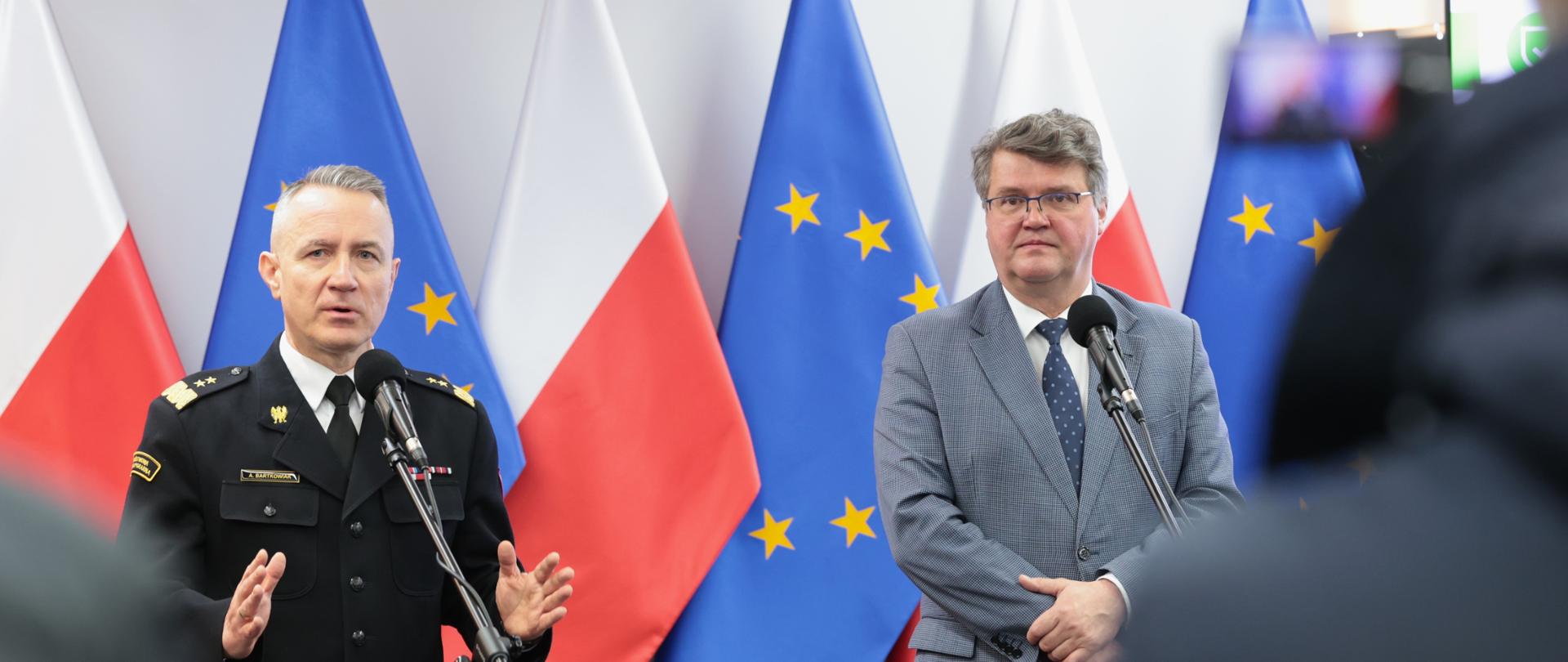 Dwóch mężczyzn stojących przed mikrofonami. Jeden z nich trzyma w dłoniach teczkę. Drugi w mundurze przemawia gestykulując. W tle widoczne są flagi Unii Europejskiej oraz Polski. 