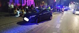 Zdjęcie wykonane w nocy. Na pierwszym planie małe osobowe auto kolory granatowego. Auto po kolizji. W tle strażacy zajmujący się kierującą i pasażerką, która odniosła obrażenia. Auto nie jest mocno uszkodzone.