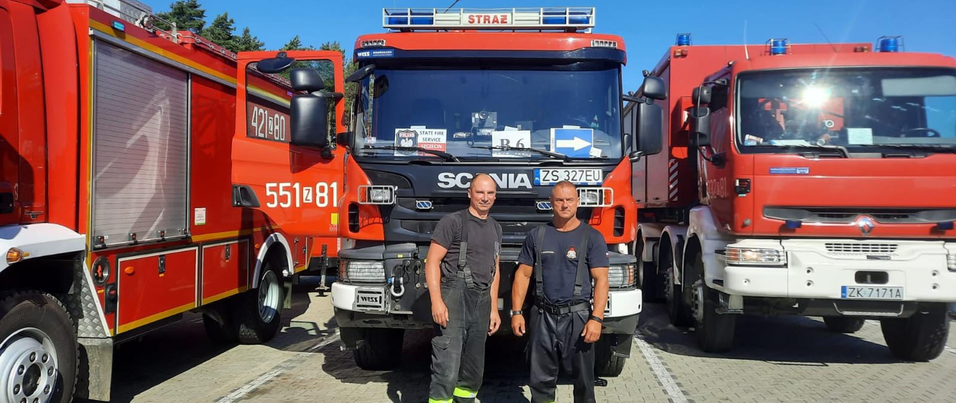 Zdjęcie przedstawia dwóch strażaków pozujących przy wozie strażackim na parkingu.