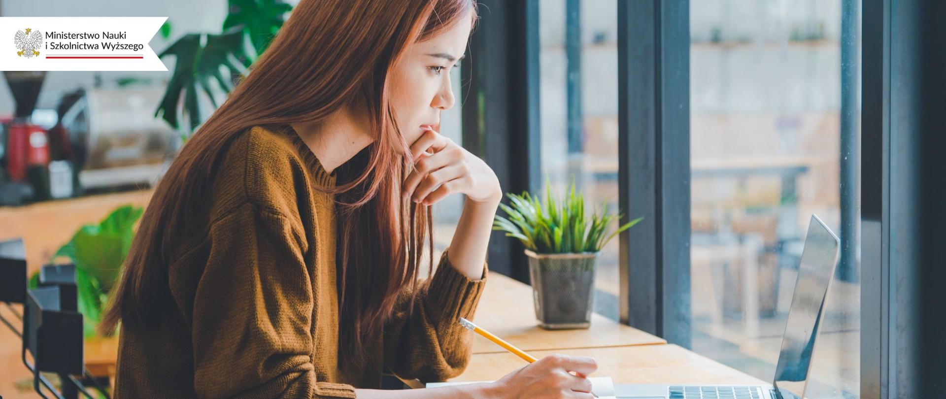 Młoda kobieta siedzi przy oknie, patrzy w otwartego laptopa.