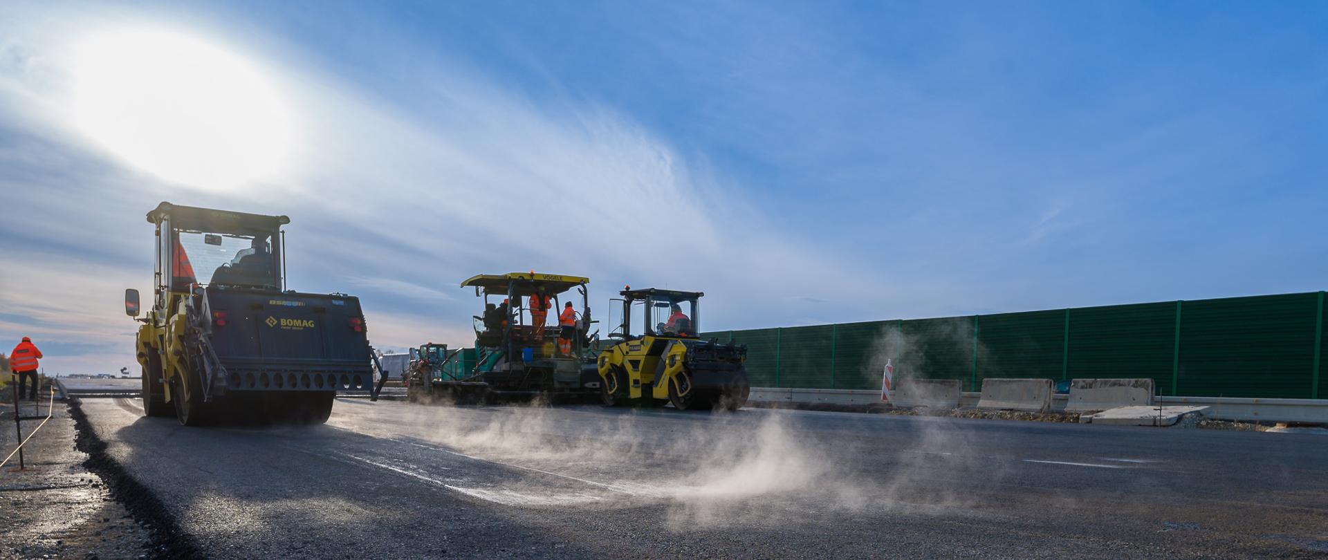 Zdjęcie przedstawia autostradę w trakcie budowy. Na pierwszym planie widoczne są maszyny drogowe, takie jak walec i rozściełacz asfaltu, które pracują nad układaniem nowej nawierzchni. W tle można dostrzec barierki ochronne oraz pracowników budowlanych ubranych w odblaskowe kamizelki. Z nawierzchni unosi się para, co sugeruje, że asfalt jest świeżo położony i jeszcze gorący.