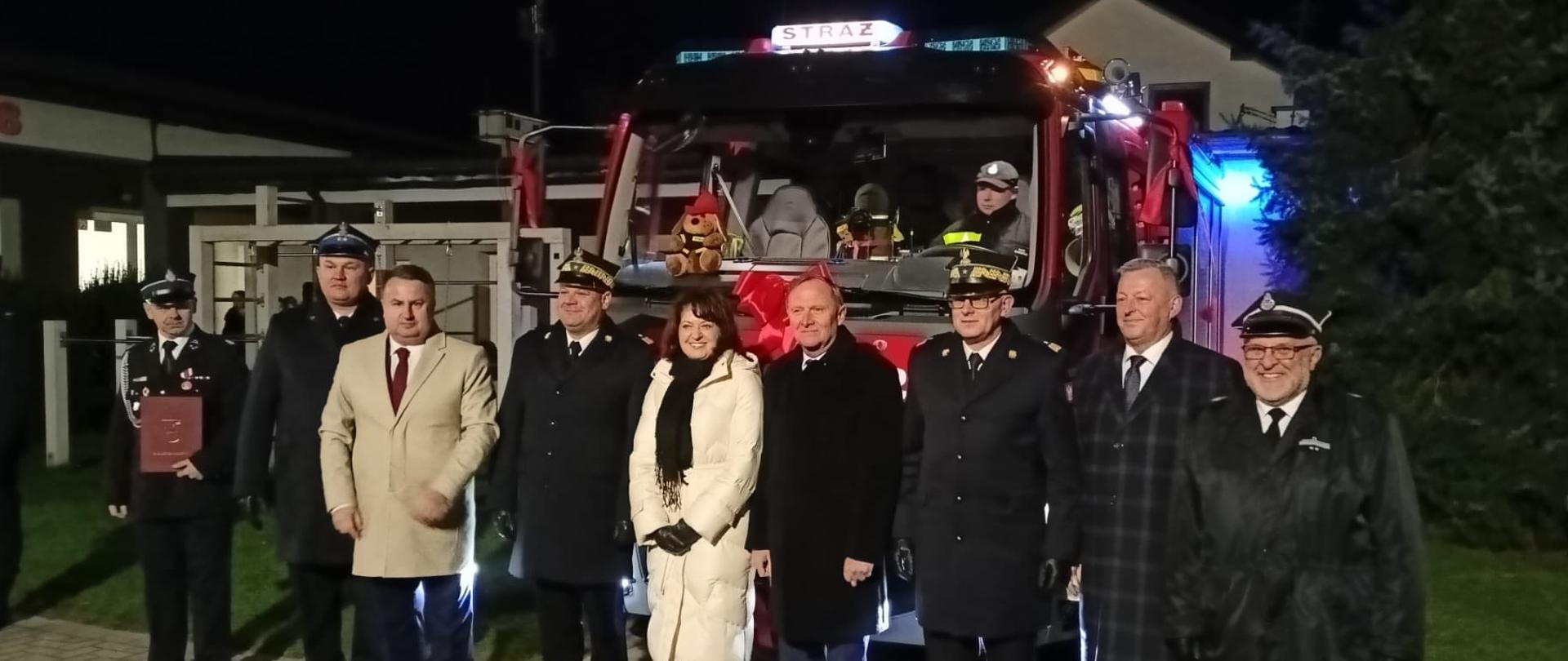 Uczestnicy uroczystości pozują przed czerwonym samochodem strażackim. 