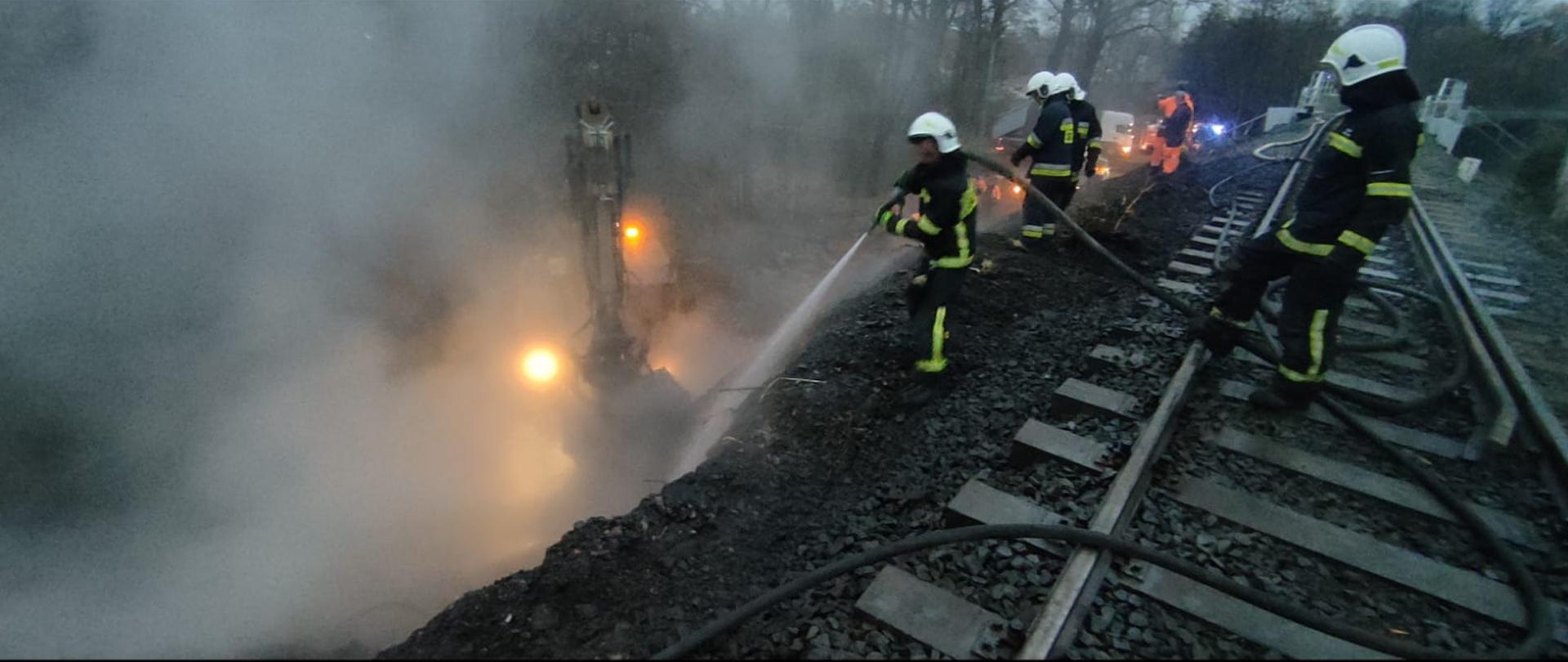 Na szczycie nasypu kolejowego stoją druhowie OSP oraz pracownicy kolei. Strażacy podają prąd wody w kierunku podstawy nasypu. W dole koparka podbiera materiał z którego wykonany jest nasyp kolejowy. W dole nasypu widać dużo białego dymu lub pary.