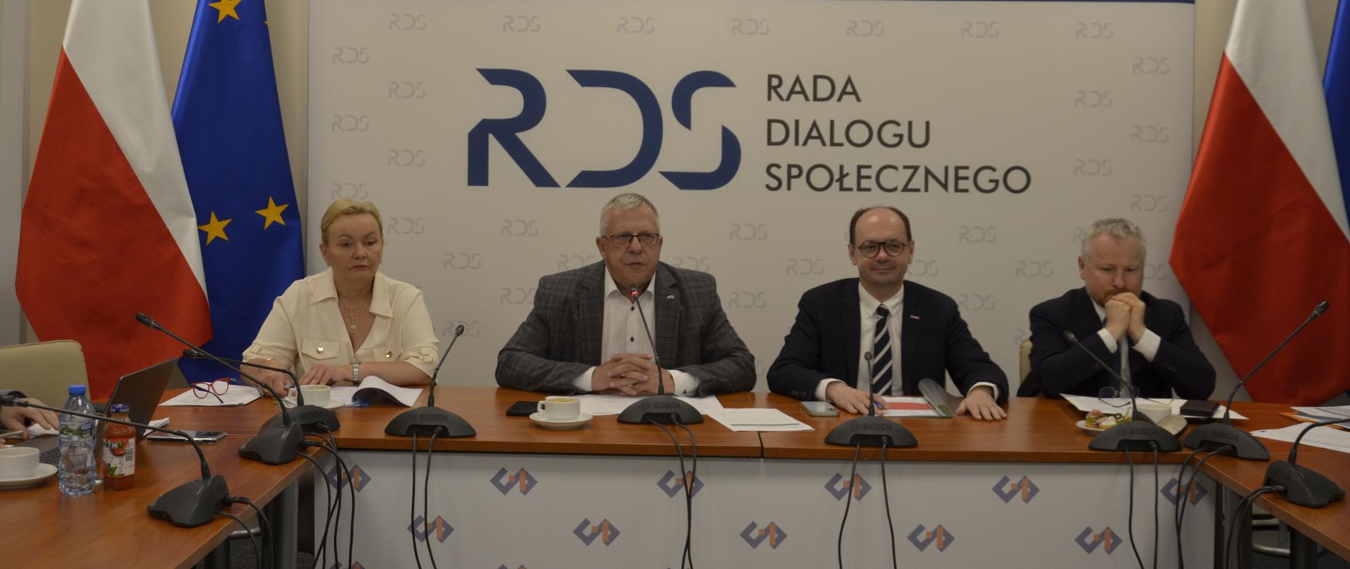 Na zdjęciu widnieje stół prezydialny za którym siedzą od lewej strony: Małgorzata Baranowska - Podsekretarz Stanu w Ministerstwie Rodziny, Pracy i Polityki Społecznej, Przewodniczący zespołu ds. budżetu, wynagrodzeń i świadczeń socjalnych – Leszek Miętek, Sebastian Gajewski - Podsekretarz Stanu w Ministerstwie Rodziny, Pracy i Polityki Społecznej Jurand Drop – Podsekretarz Stanu w Ministerstwie Finansów . Po obu stronach stołu widnieją dwie flagi: Polski i UE. W tle siedzących znajduje się baner z napisem Rada Dialogu Społecznego a nad nim wisi godło Polski.