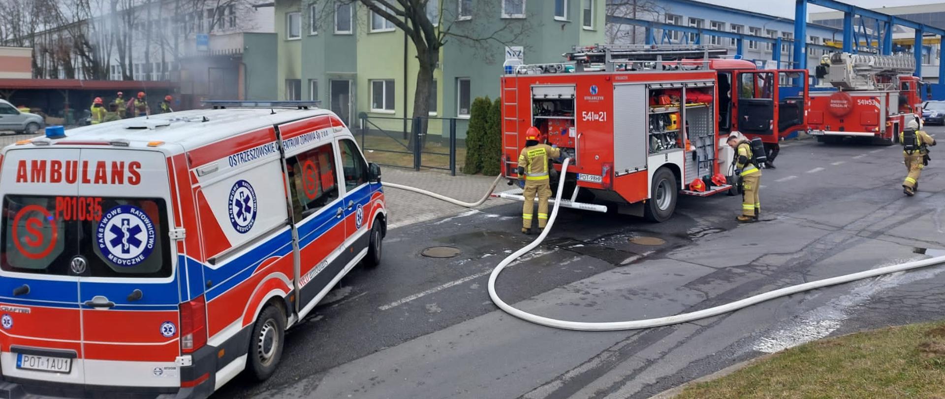 Zdjęcie przedstawia działania gaśnicze podczas pożaru budynku warsztatowego. Na zdjęciu widzimy dwa pojazdu z JRG Ostrzeszów oraz pojazd Zespołu Ratownictwa Medycznego w trakcie działań. Zdjęcie wykonane w porze dziennej przed warsztatem samochodowym.